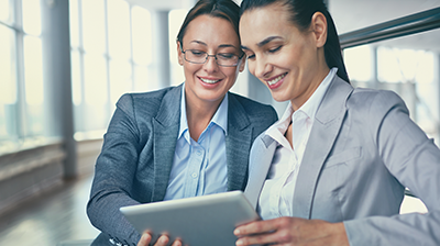 Two women in business attire looking down and smiling at tablet