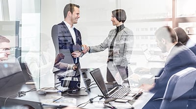 A business man and woman shaking hands in the reflection of a conference room where business people are having a meeting